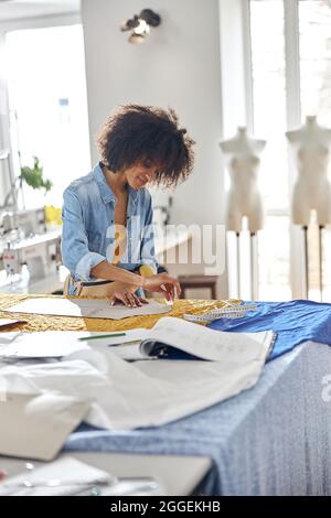 African-American dressmaker draws pattern using chalk and cardboard part in sewing workshop Stock Photo