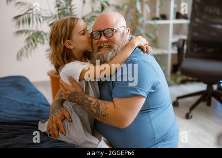 Little girl hugs and kisses smiling bearded plump father sitting on bed at home Stock Photo