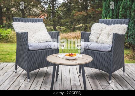 Two rattan chairs with cushions and a small table with wine glasses on a wooden terrace Stock Photo