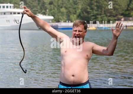Frightened man standing in water holding a poisonous snake, shouting and waving his arms. Stock Photo