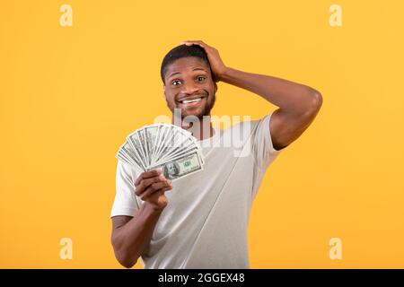 Big luck and profit. Shocked black man holding lots of money, cannot believe his big win over yellow studio background. African american guy with doll Stock Photo