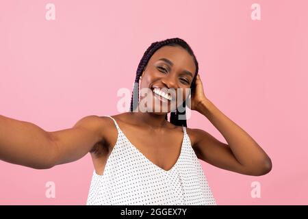 Charming young black woman with Afro braids taking selfie on pink studio background. Cheery African American lady photographing herself on smartphone, Stock Photo