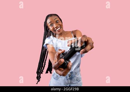 Modern entertainments concept. Excited young black lady with joystick playing video game on pink studio background. Overjoyed Afro woman holding contr Stock Photo