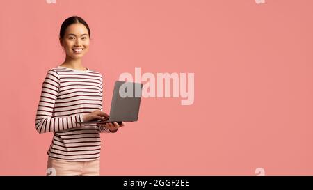 Portrait Of Young Smiling Asian Woman With Laptop Computer In Hands Standing Isolated Over Pink Background, Korean Female Working Online On Computer O Stock Photo