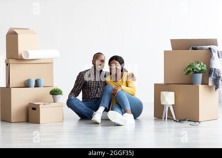 New Property. Cheerful Black Couple Sitting Among Unpacked Belongings After Moving To Their Own House, Happy African American Spouses Celebrating Buyi Stock Photo