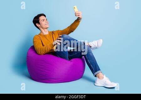 Full size photo of cool brunet young guy sit on bag do selfie wear sweater jeans isolated on blue color background Stock Photo