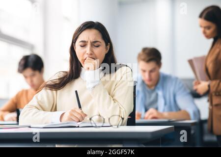 Portrait of tired female student yawning sitting at desk in classroom. Female exhausted from getting ready for test or writing coursework, feeling sle Stock Photo