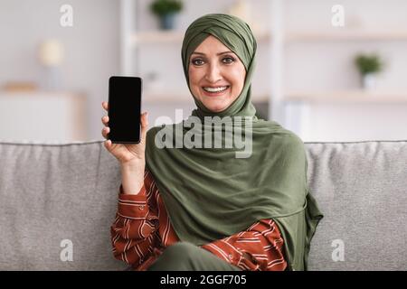 Mature Muslim Lady Showing Cellphone With Blank Screen Advertising Mobile Application Sitting On Sofa At Home, Smiling To Camera. Great New App For Yo Stock Photo