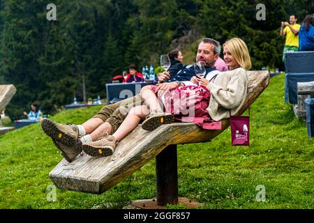 A couple relaxes with glasses of wine on a sculptural, seesaw-shaped wooden bench at the "Unplugged Taste" gourmet event on the Gompm-Alm in South Tyrol, Italy. Stock Photo