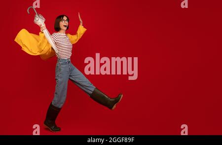 Full length shot of young woman wearing raincoat and gumboots struggling wind and walking with umbrella in hand over red studio background, panorama w Stock Photo