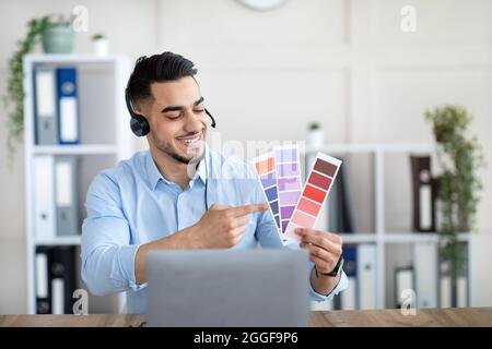 Millennial Arab guy showing color swatches at laptop webcam in office, presenting graphic design project during remote business meeting. Young male de Stock Photo