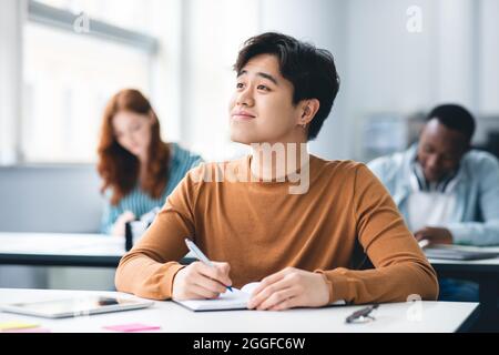 Learning Process Concept. Portrait of Smiling Asian Guy Sitting At Desk Taking Notes Writing in Exercise Copybook And Looking Aside At Window, Thinkin Stock Photo