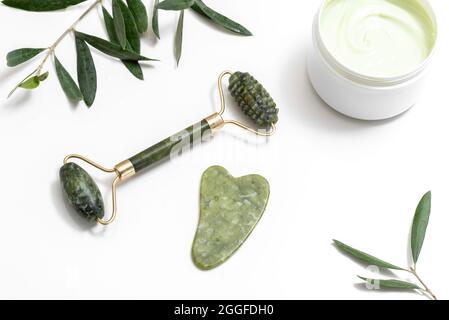 Green colored facial massage jade roller, gua sha stone, jar with olive face cream and oliva tree branches covered with drops of water on the white ta Stock Photo