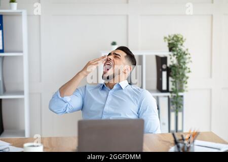 Sleepy young Arab guy yawning, tired of working with laptop at modern office. Millennial workaholic suffering from chronic fatigue, feeling sleepy or Stock Photo
