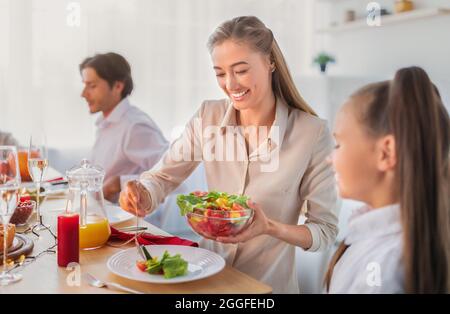 Young woman putting vegetable salad on her daughter's plate during festive family dinner at home. Little girl celebrating holiday with her parents, ha Stock Photo