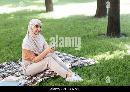 Relaxed woman using mobile phone in city park, sending text messages and chatting, watching video from social networks on free time. Happy young arab Stock Photo