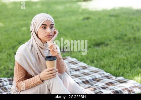 Pensive attractive millennial arabian female in hijab with cup of drink takeaway looking at empty space, sitting in park on green grass background. Co Stock Photo