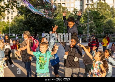 Families enjoy the street entertainment along the Southbank in London as a performer blows large bubbles across the pavement, England, United Kingdom. Stock Photo