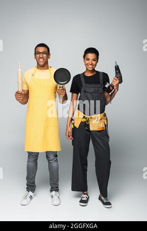 happy african american man in apron holding rolling pin and frying pan near repairwoman with electric drill on grey Stock Photo