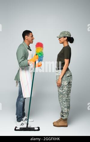 full length of female soldier in cap near cheerful male cleaner in apron with mop and dust brush on grey Stock Photo