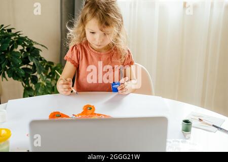 The girl spending a time drawing the paints using laptop at home. Distance learning, online education. Stock Photo