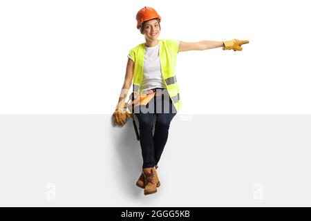 Female construction worker wearing a hardhat and a tool belt sitting on a panel and pointing isolated on white background Stock Photo