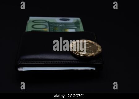 Bitcoin coin on top of wallet with  100 Euros banknotes on a dark background. Golden bitcoin on wallet one hundred euro bank notes. Stock Photo
