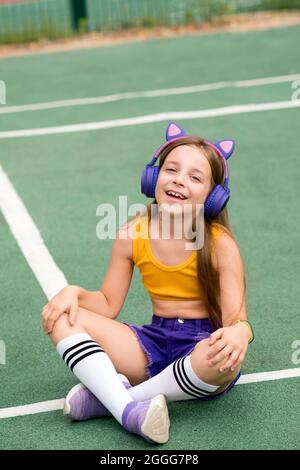 Relaxed carefree preteen girl with wireless headphones wears trendy clothes, chilling on sports ground, enjoying song single pop rock having fun alone. Soft focus Stock Photo