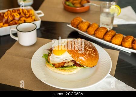 Burger with fried egg, fried bacon and melted cheese served in a tapas restaurant with other dishes such as croquettes and chorizos to hell Stock Photo