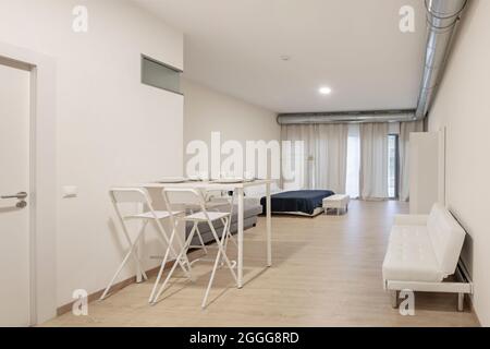 White table with breakfast service prepared with a white sofa bed in the top plan and bedroom in the back in a studio vacation rental apartment. Stock Photo