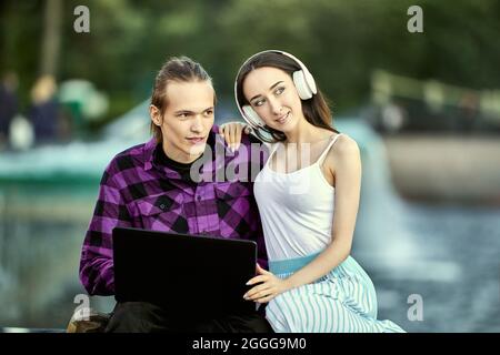 Woman in headphones and man with laptop has e-learning outdoors. Stock Photo