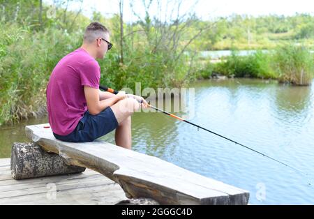 Young fisherman sitting on wooden pier, fishing in the lake. he holding the rod and looking at float Stock Photo