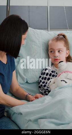 Mother explaining medication treatment to sick daughter during recovery examination in hospital ward. Hospitalized girl child resting in bed wearing oxygen nasal tube suffering disease infection Stock Photo