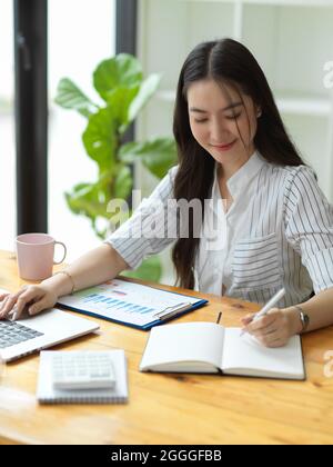 Portrait, Pretty young business entrepreneur taking notes, checking business financial on laptop computer, businesswoman working in office Stock Photo