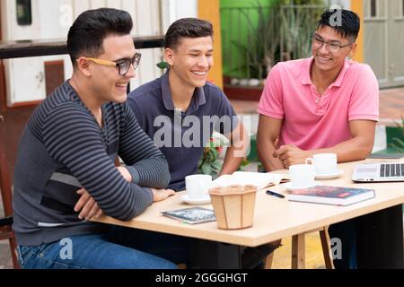 three male students sitting on benches next to a wooden desk  Stock Photo