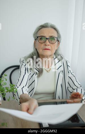 Senior grey haired businesswoman in striped jacket with eyeglasses is working with documents in here office sitting at the desk. Happy retirement Stock Photo
