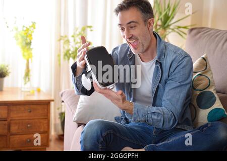 Excited and happy man looking at a virtual reality glasses sitting on a sofa in the living room at home Stock Photo