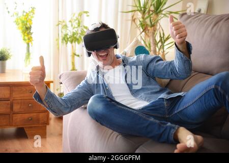 Happy entertaining man with virtual reality glasses lying on a sofa at home with ok gesture Stock Photo