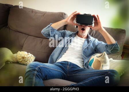Surprised man watching a movie in virtual reality glasses sitting with popcorn on the sofa at home Stock Photo