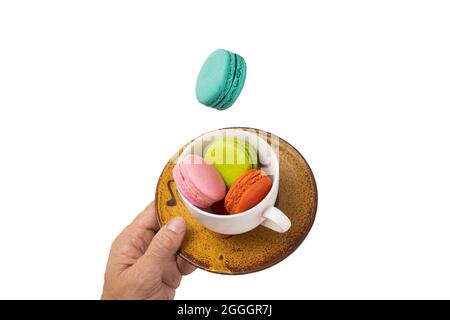 Colorful macaroons levitating on a white tea cup with a hand holding a yellowish brown saucer. Isolation on white background. Stock Photo