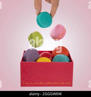 Colorful macaroons levitating on a red present  box filled with additional colorful macaroons with a hand holding one of the biscuits . Stock Photo
