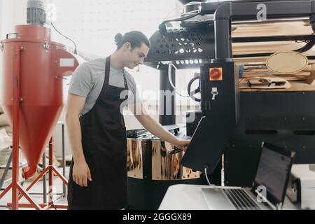 Attractive man in apron checking roasted coffee beans in cooling tray while using coffee roasting machine Stock Photo