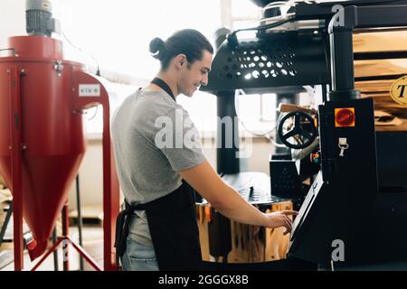 Attractive man in apron checking roasted coffee beans in cooling tray while using coffee roasting machine Stock Photo
