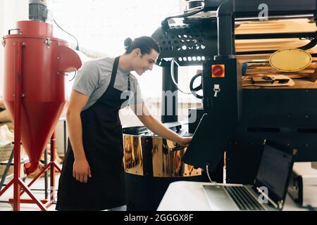 Attractive man in apron checking roasted coffee beans in cooling tray while using coffee roasting machine Stock Photo