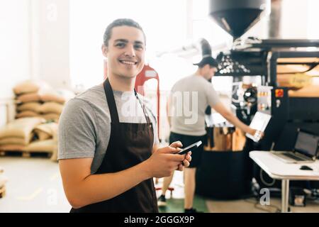 Portrait of handsome modern barista wearing apron use phone while standing at coffee roasting machine in local roastery Stock Photo