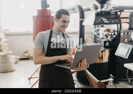 Young man working at coffee production. Barista adjusting coffee machine and using special application on his laptop while controlling coffee grounds Stock Photo