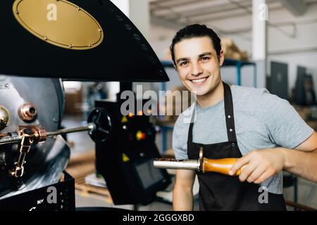 Handsome man in apron looking at freshly roasted coffee beans while standing by coffee roasting machine. Stock Photo