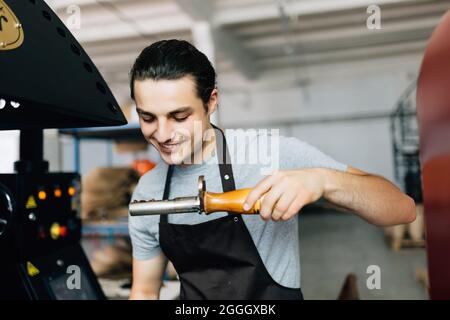 Handsome man in apron looking at freshly roasted coffee beans while standing by coffee roasting machine. Stock Photo