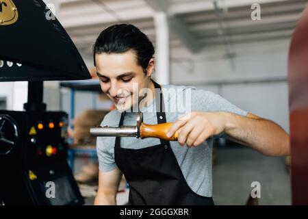 Handsome man in apron looking at freshly roasted coffee beans while standing by coffee roasting machine. Stock Photo
