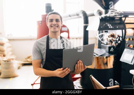 Young man working at coffee production. Barista adjusting coffee machine and using special application on his laptop while controlling coffee grounds Stock Photo